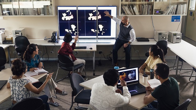 A mature man can be seen pointing to scientific matters on three large screens at the front of a classroom. There are six mature students in the room watching the presentation.